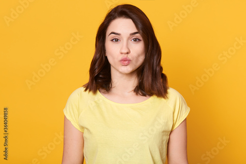 Portrait of lovely brunette female rounds lips as going to get kiss from boyfriend, looks at camera with pleased expression, isolated over yellow background.