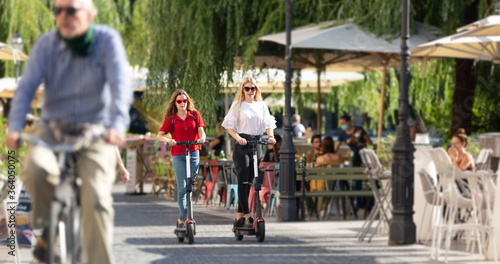 Trendy fashinable teenager girls riding public rental electric scooters in urban city environment. New eco-friendly modern public city transport in Ljubljana, Slovenia.