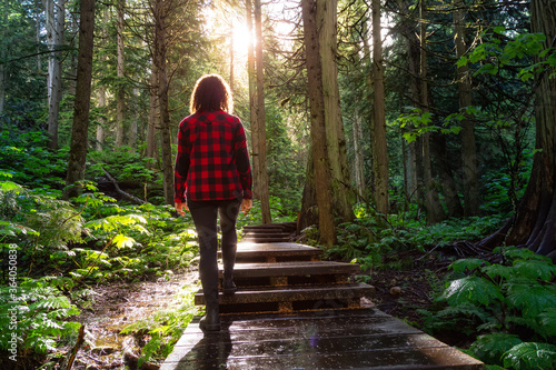 Adventure Girl Walking on a Wooden Pathway in the Rain Forest during a vibrant sunny day. Taken on Giant Cedars Boardwalk Trail in Mt Revelstoke National Park, British Columbia, Canada. photo