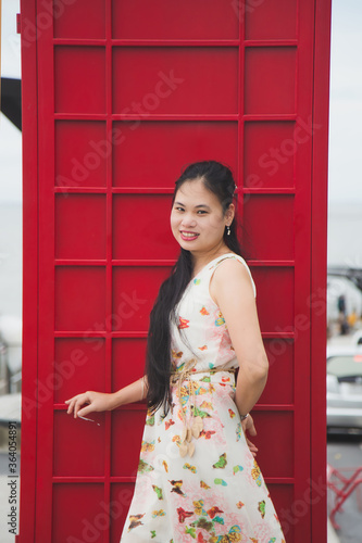 Close up portrait long hair Asian woman in a sweet beige long dress standing next to a red telephone booth with nature background.