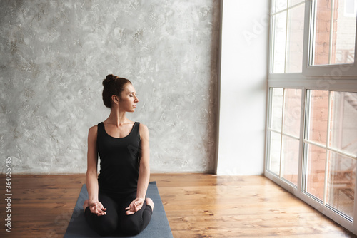 A middle-aged woman practicing yoga, doing Sukhasana exercise, Easy Seat pose, wearing black sportswear at home or yoga studio with big window background. Mindfulness and healthy lifestyle concept.