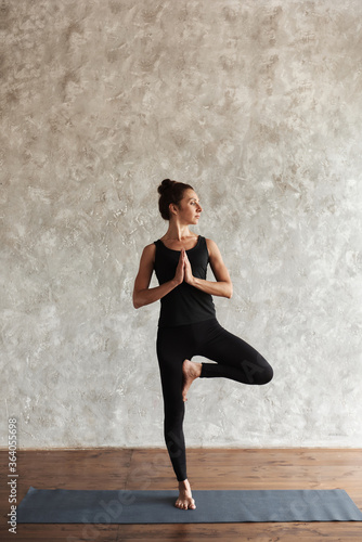 Strong middle-aged woman practicing yoga, looking away concentrated, doing Vrksasana exercise, Tree pose, working out, wearing black sportswear at yoga studio with gray wall. Healthy life concept.