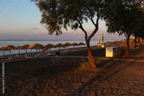 Wallpaper Mural Santorini black beach Perissa, Perivolos illuminated by the morning rays of the sun. Beach chairs with umbrellas. Two olive trees. Beautiful calm sea. Torontodigital.ca