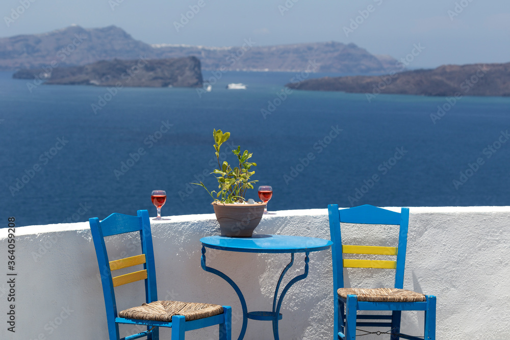 Santorini, a romantic seating area overlooking the Caldera, two blue chairs and a blue table by a white wall with a semicircular framed blue window overlooking the beautiful blue sea.