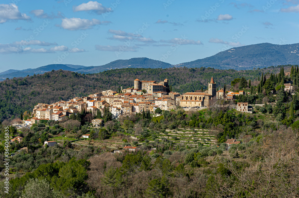 Provencal village in the hinterland. Perched on a hill, the village has a church with a bell tower with a glazed tile roof. It is dominated by a castle with a round tower. Some white clouds in the blu