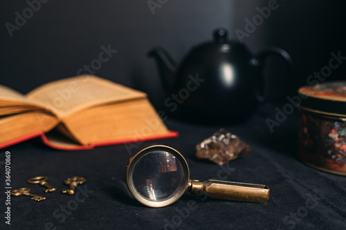 Collection of mysterious items: old book, magnifying glass, little keys, black tea pot, stone and tin box. Black background.