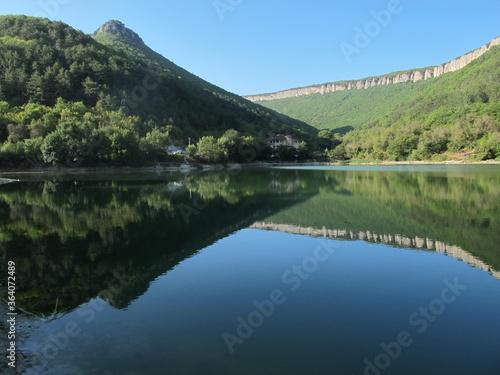 The panorama of a mountain lake at the foot of a mountain plateau, the mirror surface of the water surface reflects high mountains and rocks, overgrown with dense green forest on a clear summer day. 