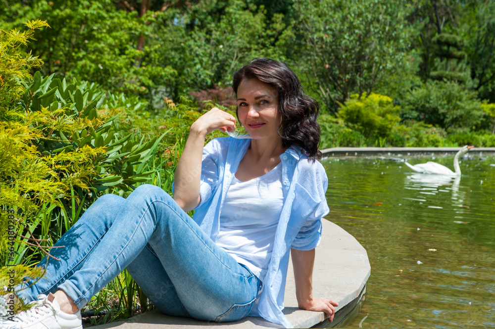 Fototapeta premium .Young brunette woman posing on a wooden pier near the lake. Girl dressed in jeans and t-shirt smiling. Swan lake, outdoor.