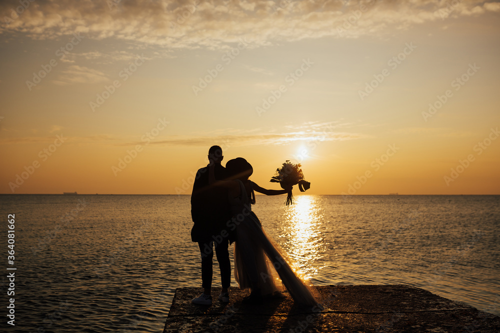 Silhouette of a loving couple on a background of evening sunny sky, sunset. Young wedding couple stands at the sea beach looking at sunset.  Copy space.