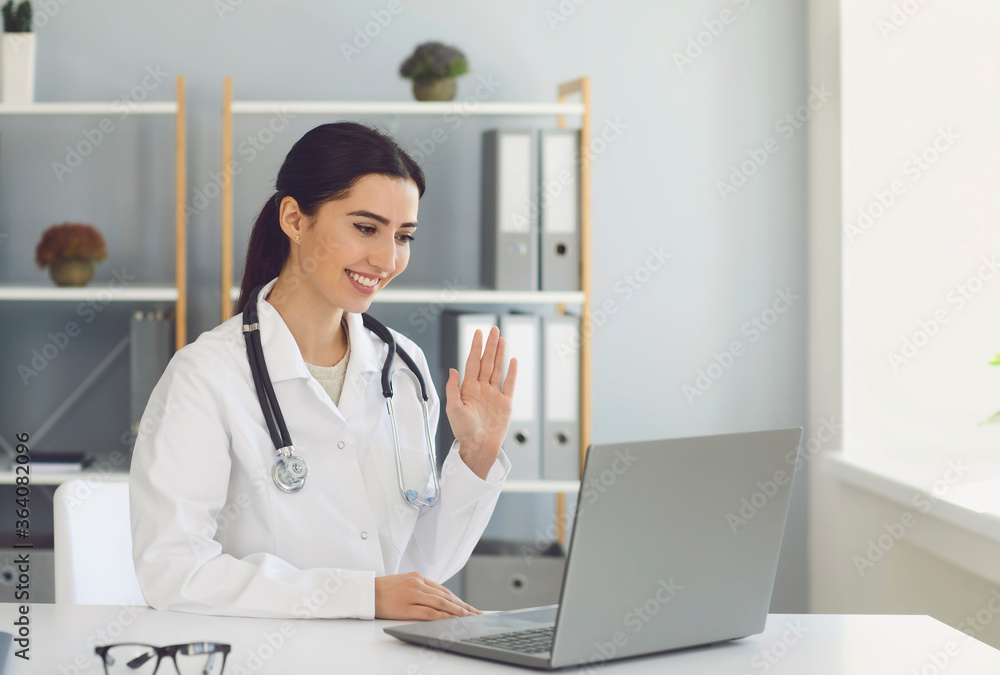 Hispanic female doctor waving to a patient has a video conference laptop sitting at a table in a clinic office. Hispanic female doctor waving to a patient has a video conference laptop sitting at a table in a clinic office.
