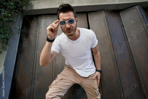 Stylish handsome young man with a bag near the metal gate, wearing white t-shirt with sunglasses 