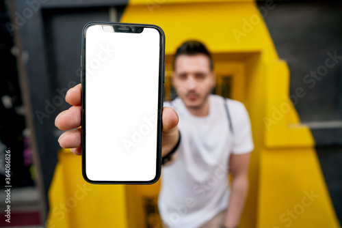 Hipster guy showing cellphone device with copy space area for internet advertising, blurred millennial male  promote mock up mobile phone with blank screen for your website, face out of focus.