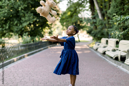 Portrait of happy little African girl child in green city park throwing up teddy bear toy, flying in the air. Smiling dark skinned kid girl in blue dress playing in summer park.