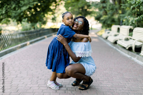 Fototapeta Naklejka Na Ścianę i Meble -  Full length portrait of smiling young happy african mother hugging her little baby daughter with tenderness and love standing in the park. Mother and daughter in blue dresses walking in park