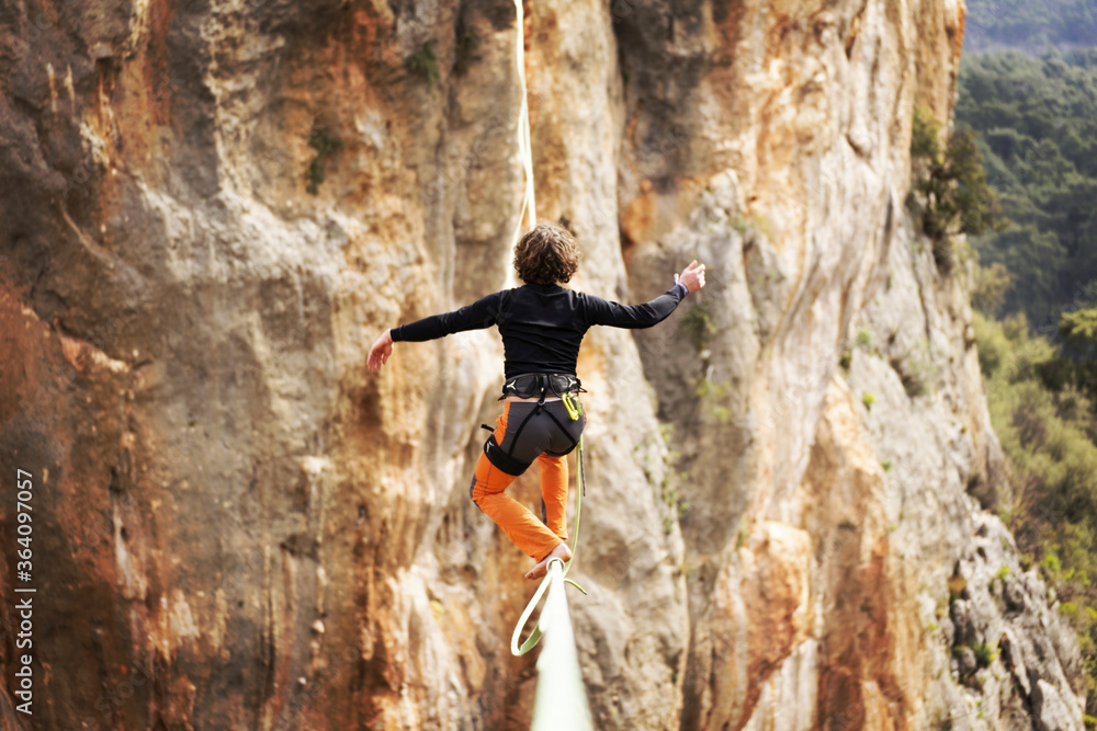 Fototapeta premium A man is walking along a stretched sling. Highline in the mountains. Man catches balance. Performance of a tightrope walker in nature. Highliner on the background of valley.