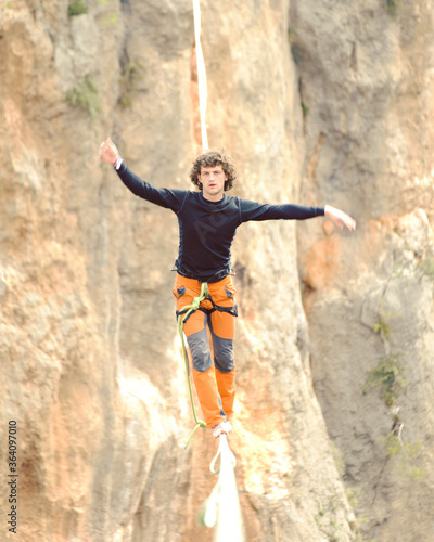 A man is walking along a stretched sling. Highline in the mountains. Man catches balance. Performance of a tightrope walker in nature. Highliner on the background of valley. © vetal1983