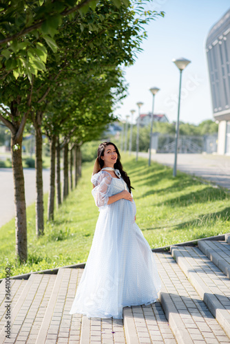 Pregnant woman posing in a blue dress on a background of green trees.