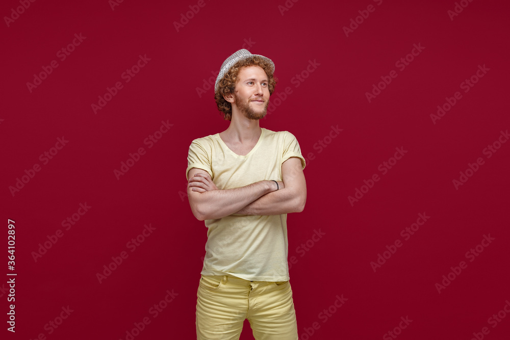 Fashionable hipster man posing with his arms crossed in studio. Smiling man in straw hat looking away from camera isolated in red background.