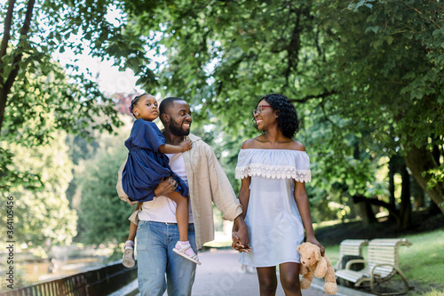 happy young african family spending time outdoor in park on a summer day. Joyful smiling couple holding hands and walking down the park alley, man holds cute little daughter in blue dress