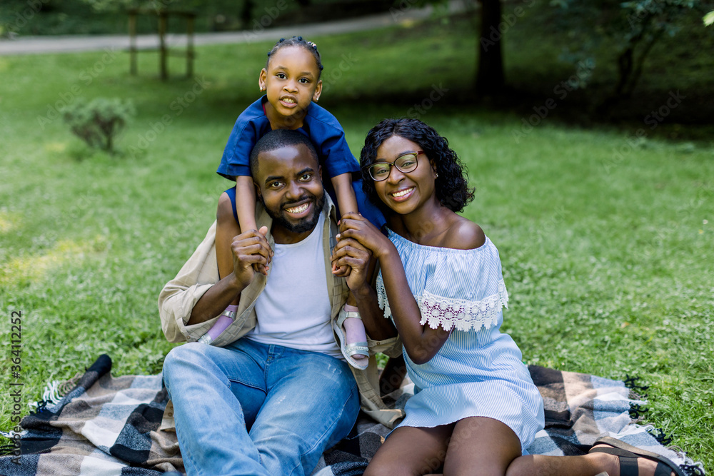Adorable young African family with cute little daughter, having their weekend time in a city park. Image of cheerful family looking at the camera while picnicking in the park