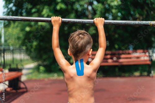 The young athlete pulls himself on the horizontal bar. Doing sports in the park. Teip on the back and neck, curvature of the spine, scoliosis, back problems