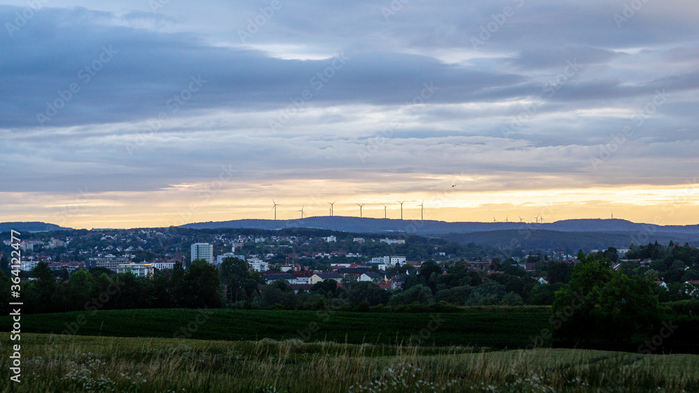 Helikopter fliegt am Horizont entlang einer Reihe von Windrädern bei orangenfarbenem Sonnenuntergang  über die Stadt Bayreuth in Oberfranken, Bayern, Deutschland.