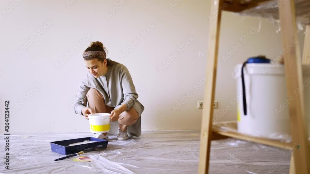 smiling lady pours white paint from plastic bucket into blue container and matches with templates and tools on brown ladder on foreground in new flat smiling lady pours white paint from plastic bucket into blue container and matches with templates and tools on brown ladder on foreground in new flat