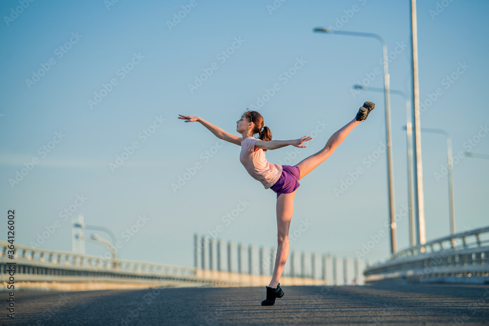 Girl gymnast engaged in the summer on the street, on a background of blue sky, twine, stretching, arabesque Girl gymnast engaged in the summer on the street, on a background of blue sky, twine, stretching, arabesque