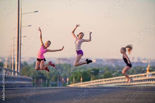 Three slender sports girls jumping on the road on a summer evening against the backdrop of the city panorama and pink sunset sky