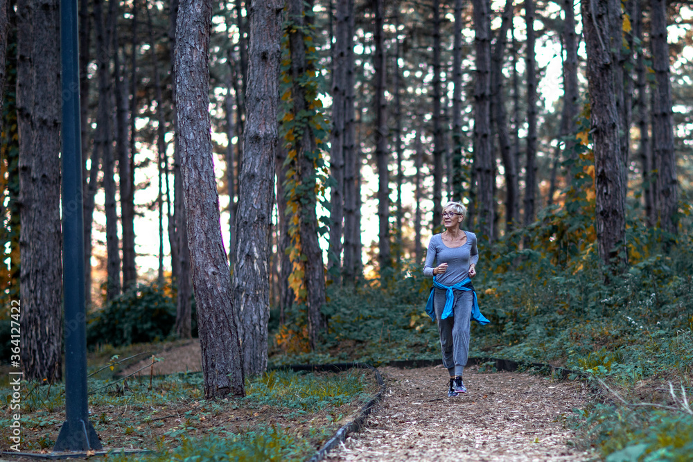 Aged woman jogging in park between woods at the end of day and smile