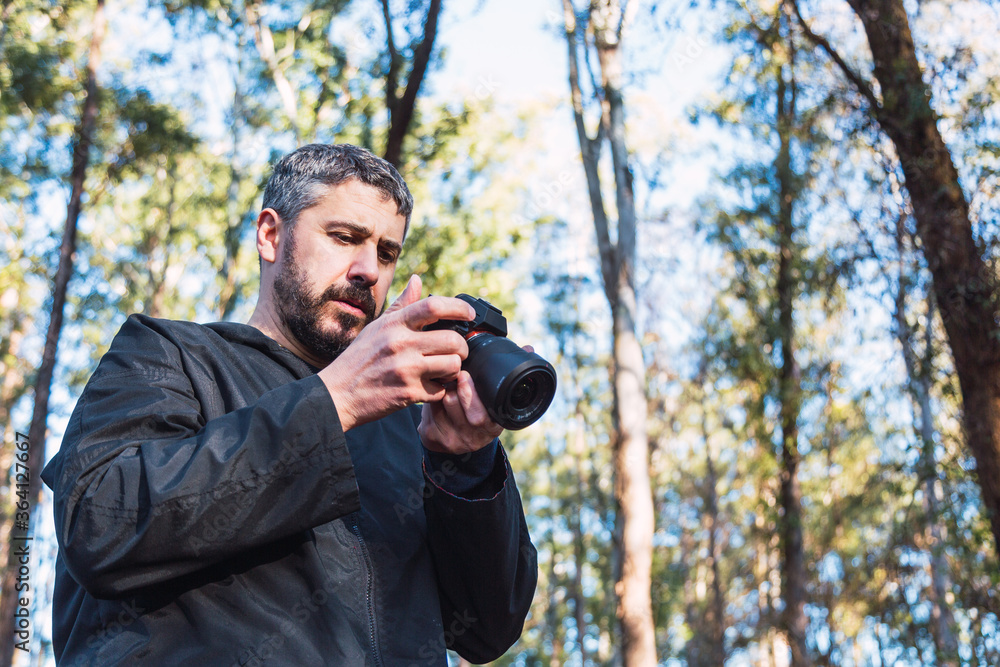 A man checks his camera. He has a beard, and gray hair, about 40 years old, and walks in a park with many trees in the background. A man checks his camera. He has a beard, and gray hair, about 40 years old, and walks in a park with many trees in the background.