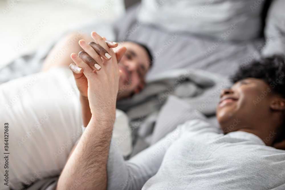 Young multiethnic couple holding hands while lying in bed in morning Young multiethnic couple holding hands while lying in bed in morning