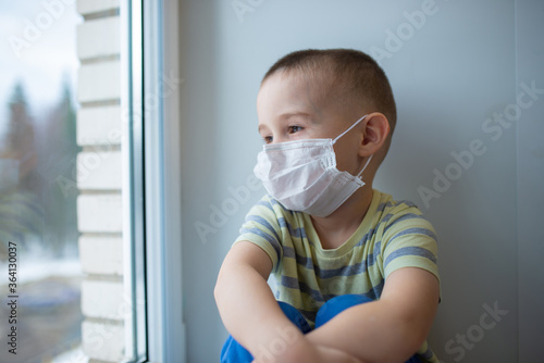 Sad little boy in medicine mask sitting near window and looking outside, quarantine at home, virus protection
