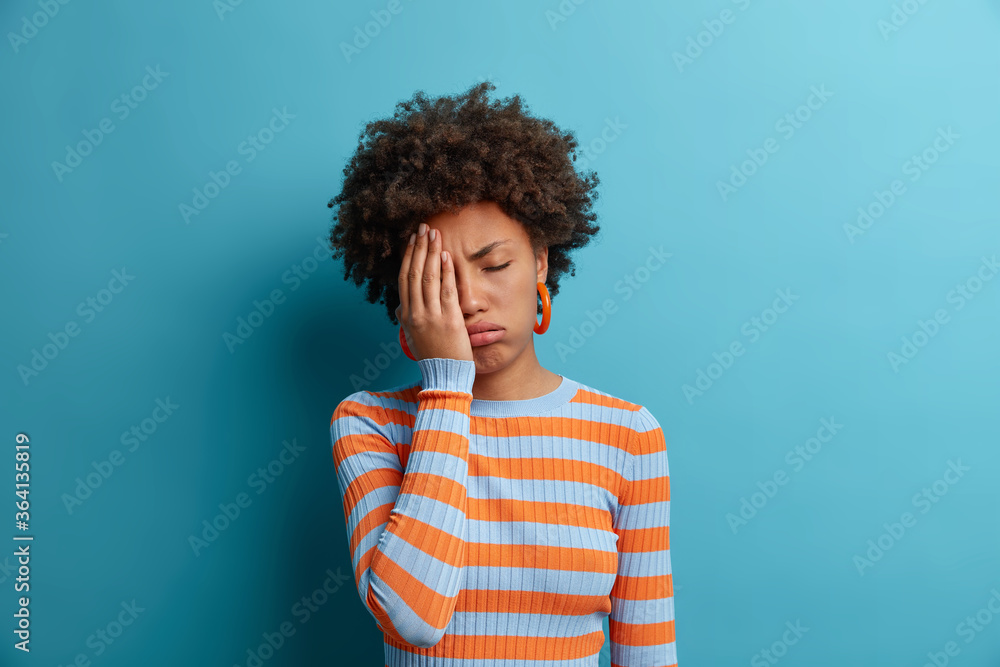 Displeased exhausted woman makes face palm, cannot work any more, closes eyes and feels bad, wears casual striped jumper, wakes up very early, isolated on blue background, suffers from headache Displeased exhausted woman makes face palm, cannot work any more, closes eyes and feels bad, wears casual striped jumper, wakes up very early, isolated on blue background, suffers from headache