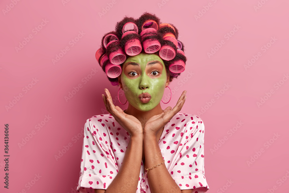 Magnetic ethnic woman keeps lips rounded, spreads palms over face, dressed in nightwear, poses with hair curlers and beauty mask for healthy skin and grooming compexion, isolated on pink background