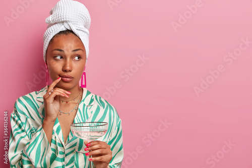 Thoughful lady enjoys drinking martini cocktail after taking shower, looks aside, has well cared complexion, healthy skin, wears dressing gown and towel, isolated on pink studio wall, blank space