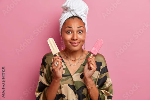 Indoor shot of pleased young dark skinned woman eats delicious appetizing ice cream of vanilla and strawberry flavor, enjoys summer dessert, dressed in casual domestic clothes, poses indoor.