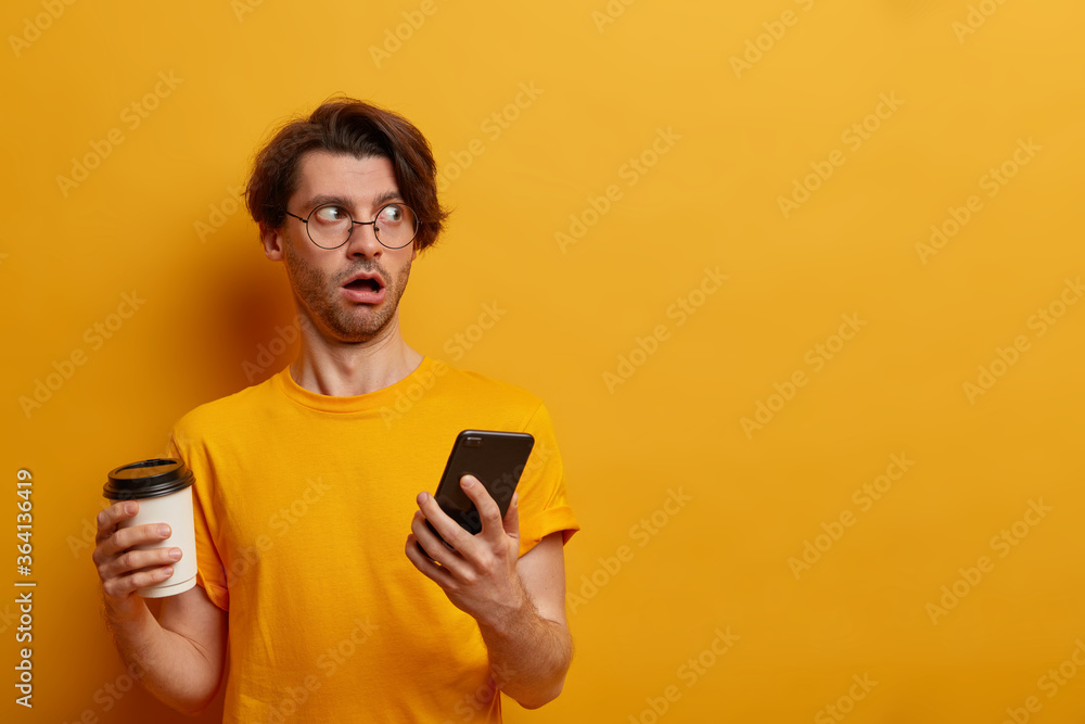 Impressed speechless guy looks aside with astounded expression, uses mobile phone and takeaway coffee, reads awesome news, chats with friend, wears round spectacles and yellow t shirt, blank space Impressed speechless guy looks aside with astounded expression, uses mobile phone and takeaway coffee, reads awesome news, chats with friend, wears round spectacles and yellow t shirt, blank space