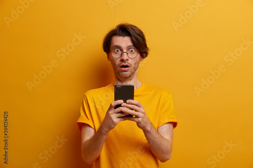 Stupefied man distracted from screen of smartphone, watches horrible video, gasps from wonder, got unexpected message, wears round sprectacles and t shirt, isolated over vivid yellow background