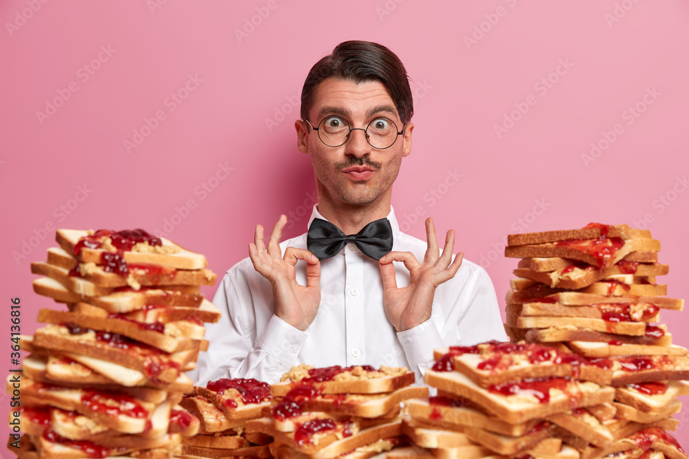 Funny elegant man employee fixes bowtie, ready for first working day in restaurant, wears classic formal clothing and round glasses, poses near table full of bread toasts isolated over pink background Funny elegant man employee fixes bowtie, ready for first working day in restaurant, wears classic formal clothing and round glasses, poses near table full of bread toasts isolated over pink background