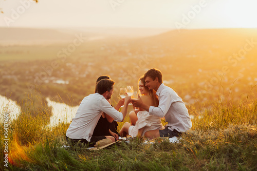 Couples having a picnic in the countryside. People with glasses of white wine on the background of river at the picnic. Cute couples looking at each other against the backdrop of the river and sunset.