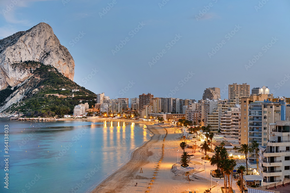 Fototapeta premium Panoramic view from above of Calpe cityscape. Street lights illuminating seafront promenade during sunset. Empty sandy beach and rocky mountain of Penon de Ifach. Costa Blanca Alicante province, Spain