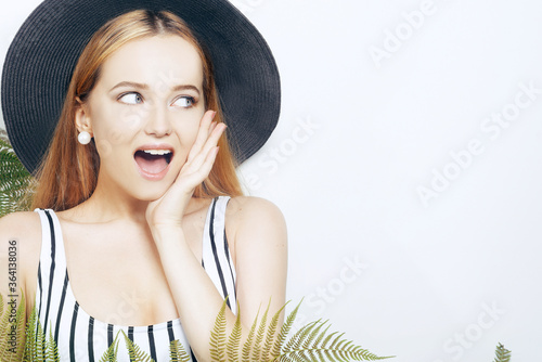 The girl in the tropical leaves in a swimsuit in a big black hat looks away shocked, surprised. Space for text, empty space. Model summer photo.
