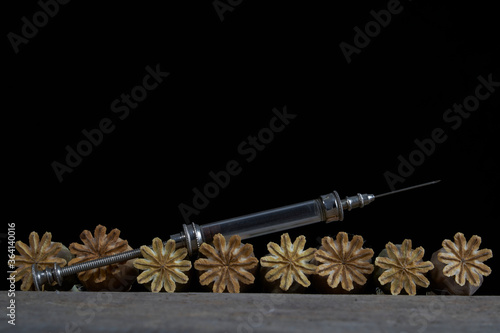 Glass syringe lies on dried opium poppy capsules on a wooden table against a black background