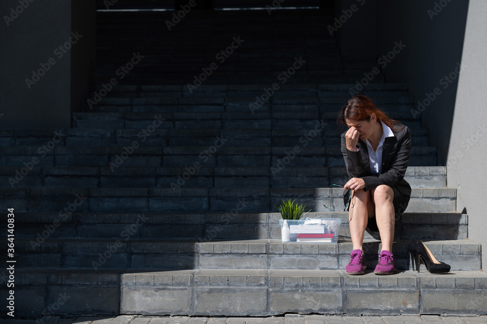 Depressed woman dressed in medical mask is fired and is sitting on the stairs with a box of personal belongings. Female office worker in suit and sneakers outdoors. Unemployment in the economic crisis Depressed woman dressed in medical mask is fired and is sitting on the stairs with a box of personal belongings. Female office worker in suit and sneakers outdoors. Unemployment in the economic crisis