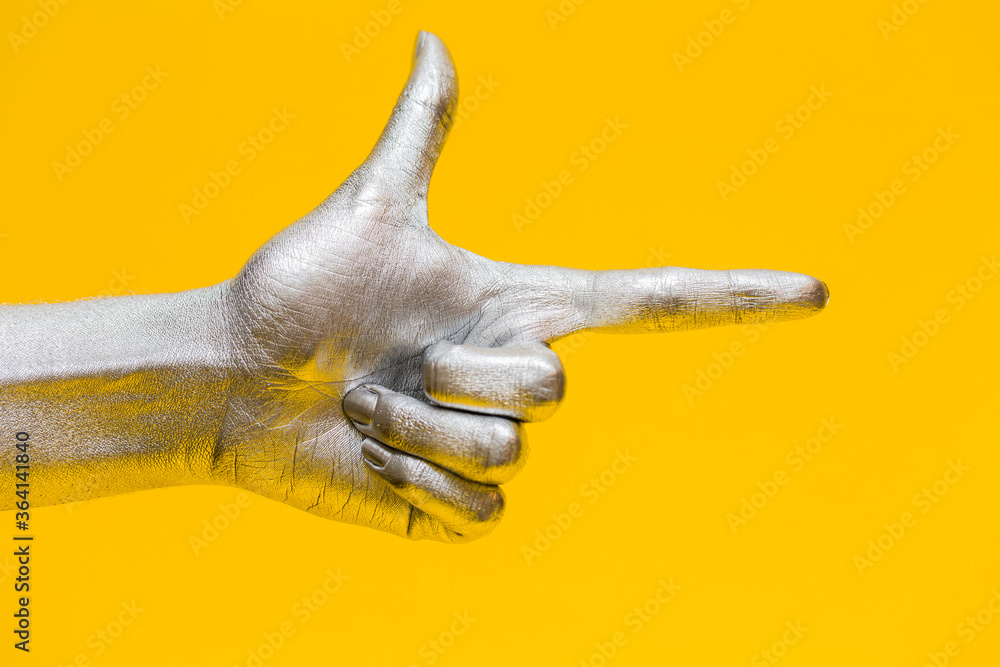 Fototapeta premium Elegant female hand with a silver paint on it shows gesture bang bang isolated on a yellow background. Free space for text