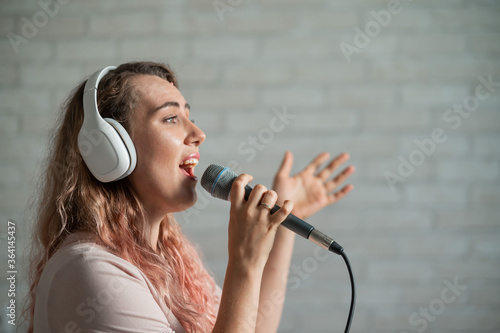 Close-up portrait of a caucasian woman with curly hair singing into a microphone. Beautiful emotional girl in white headphones sings a song in home karaoke and actively gestures against a brick wall.