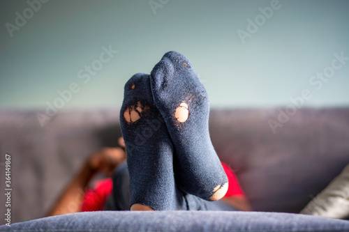 Bottom of old worn out socks with holes in it. Man in living room with feet propped up on ottoman, relaxing on the couch at home. Concept of poverty