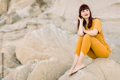 Summer lifestyle fashion image of pretty woman wearing stylish yellow overalls, sitting on the sand stone in quarry, enjoying moment and looking at camera.