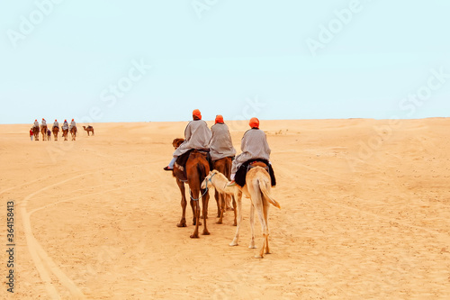 Tourists dressed in striped robes and orange turbans on their heads riding on two-humped camels for a walk in the Sahara desert, Tunisia. View from the back